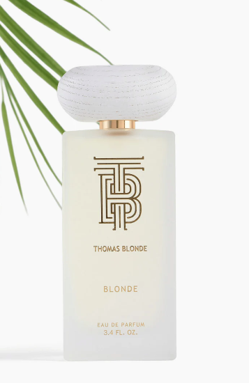 Thomas Blonde Blonde Eau de Parfum bottle with a frosted glass body, gold lettering, and a textured white cap, styled with green palm leaves on a white background.