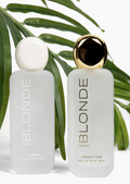 Two frosted glass bottles of Thomas Blonde Joshua Tree Hair & Body Mist, one with a white cap and one with a gold cap, styled with green palm leaves on a white background.