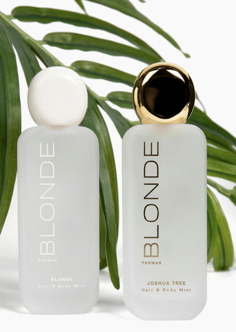 Two frosted glass bottles of Thomas Blonde Joshua Tree Hair & Body Mist, one with a white cap and one with a gold cap, styled with green palm leaves on a white background.