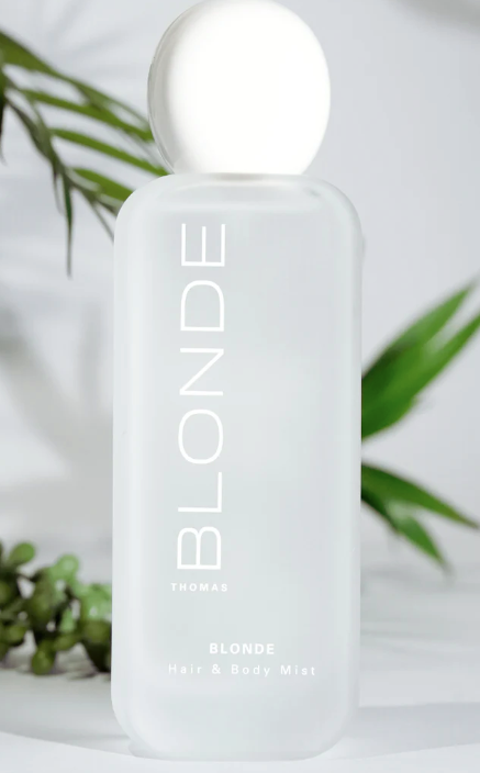 A frosted white bottle of Thomas Blonde Hair & Body Mist with a matching spherical cap, set against a soft white background with blurred green leaves.