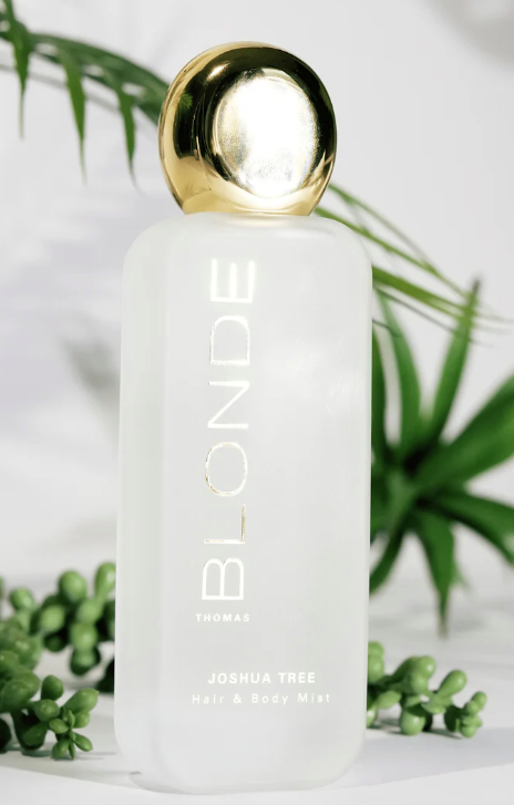 A frosted white bottle of THOMAS BLONDE Joshua Tree Hair & Body Mist with a metallic gold spherical cap, styled with green plants on a white background.