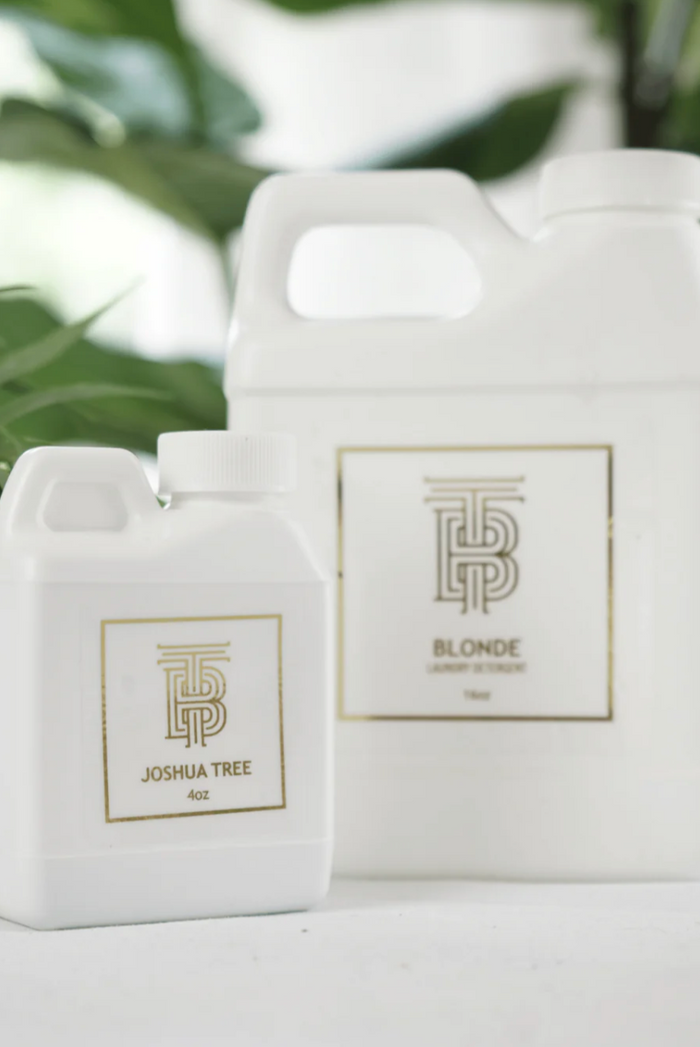 Two white plastic bottles of hair color, one smaller bottle labeled 'JOSHUA TREE Acid' and one larger jug labeled 'BLONDE', with a gold monogram logo on the front of each bottle.