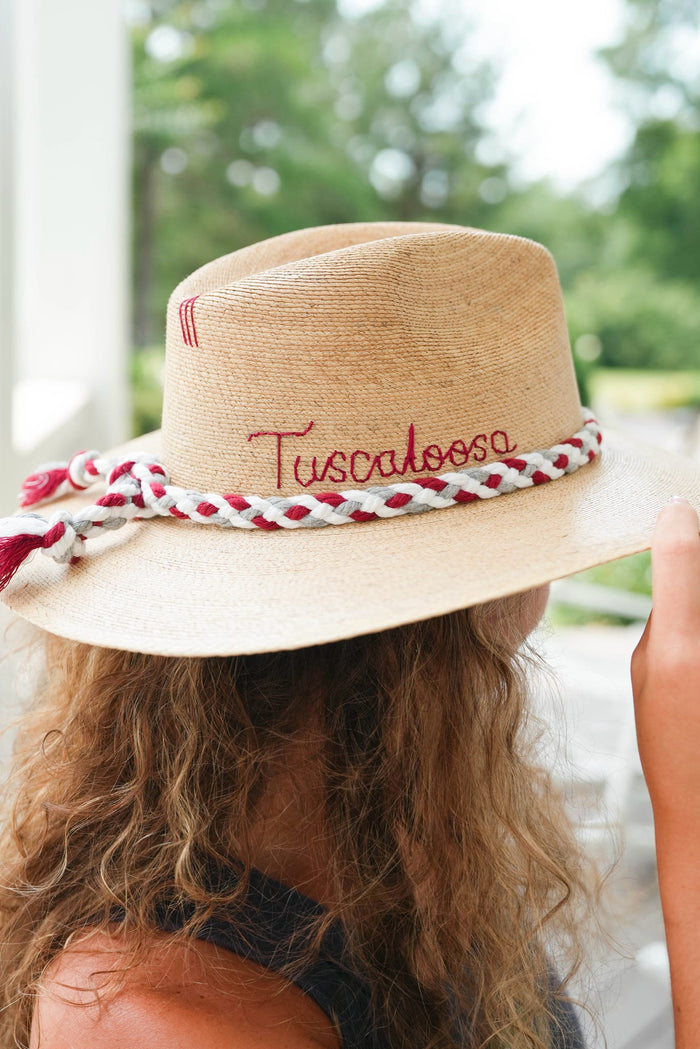 A straw fedora hat with 'Tuscaloosa' embroidered on the crown and a red-and-white braided rope band, shown on a person outdoors.