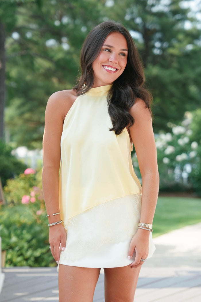 Woman wearing a yellow and white dress standing outdoors with greenery in the background