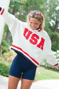 Woman wearing a white 'USA' sweatshirt with red and blue accents outdoors.