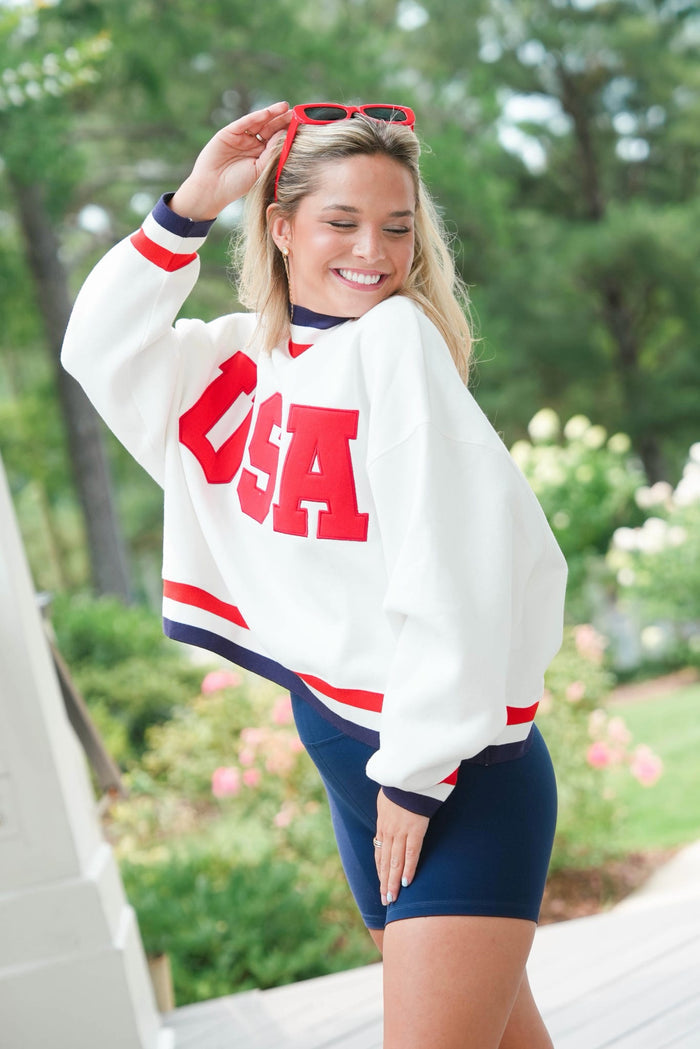 Woman wearing a white 'USA' sweatshirt with red and blue accents outdoors.