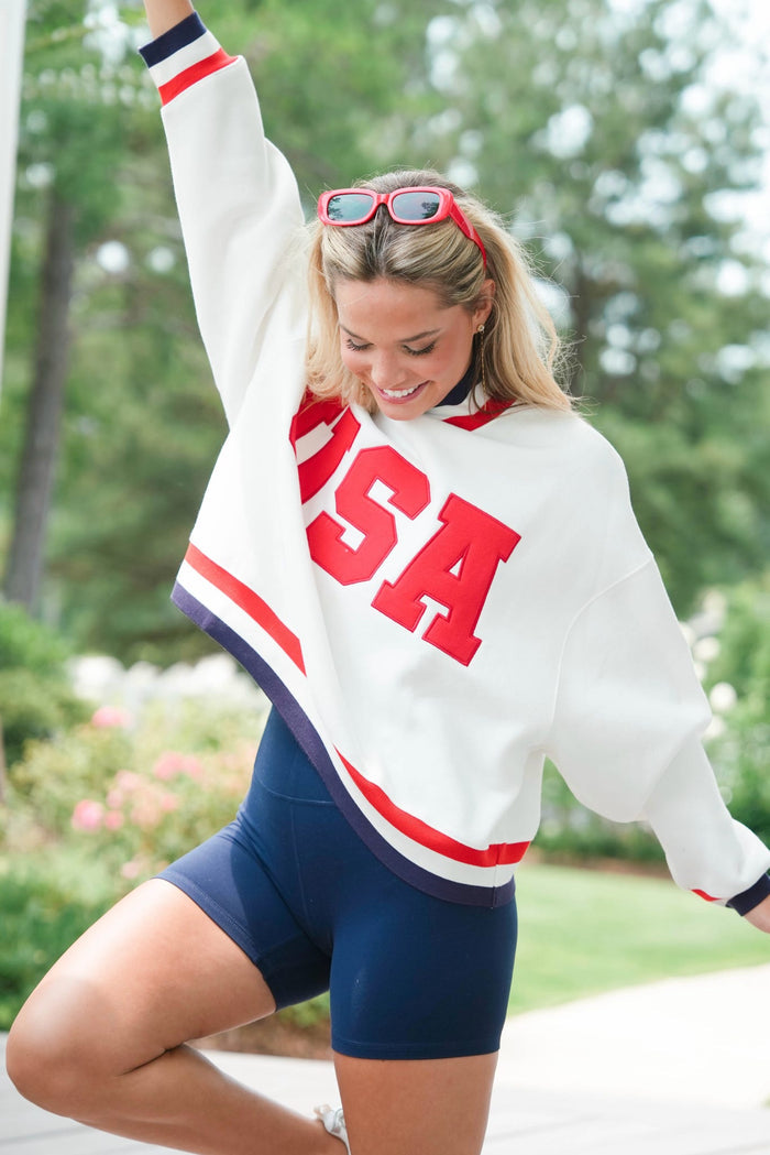 Woman wearing a white 'USA' sweatshirt with red and blue accents outdoors.