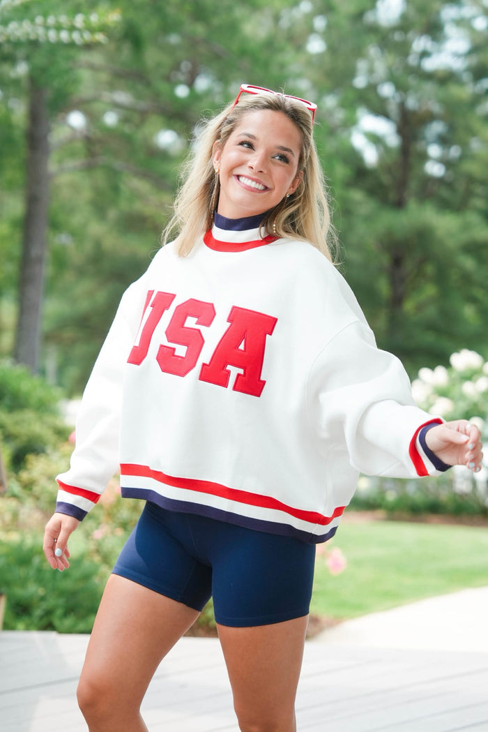 Woman wearing a white 'USA' sweatshirt with red and blue accents outdoors.