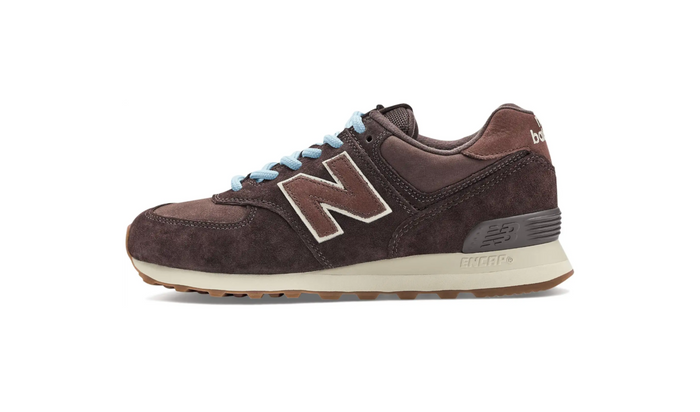 A New Balance 574 sneaker in dark brown suede and leather, featuring light blue laces, a large white and brown 'N' logo on the side, and a cream-colored midsole.