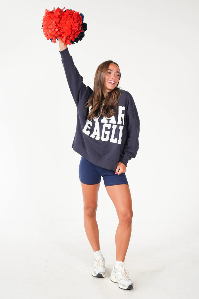 A woman modeling a navy blue sweatshirt with 'EAGLE' printed in white, paired with navy shorts and white sneakers, holding a red and black pom-pom against a plain white background.