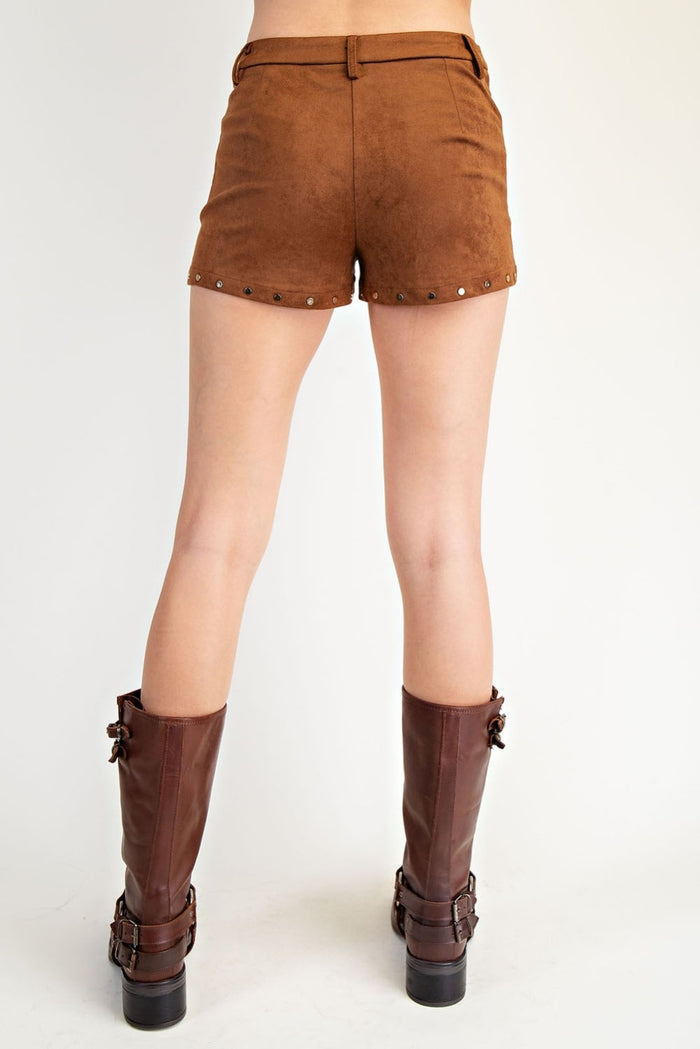 Brown suede shorts with metal studs along the hem, shown on a model from the back wearing dark brown buckle-strap boots against a white studio background.