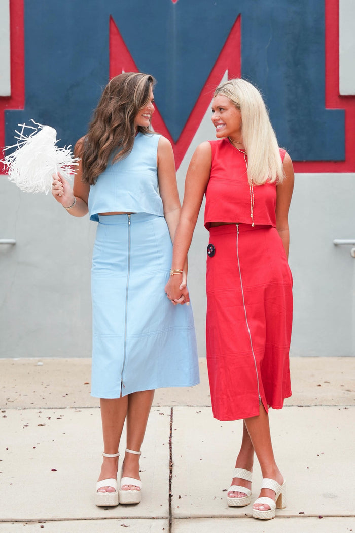 Two women standing outdoors and holding hands, wearing matching two-piece outfits. One outfit is light blue and the other is red, both with sleeveless cropped tops and midi skirts.