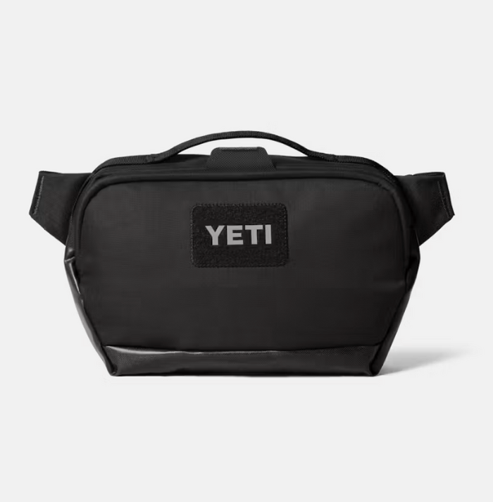 A black YETI shoulder bag with a top handle and a black rectangular patch on the front featuring the white YETI logo, set against a light gray background.