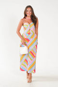 A woman modeling a sleeveless, spaghetti-strap midi dress with a colorful pastel geometric stripe pattern in pink, yellow, blue, green, and orange. She is holding a small white handbag and wearing a pink beaded necklace, set against a plain white studio background.
