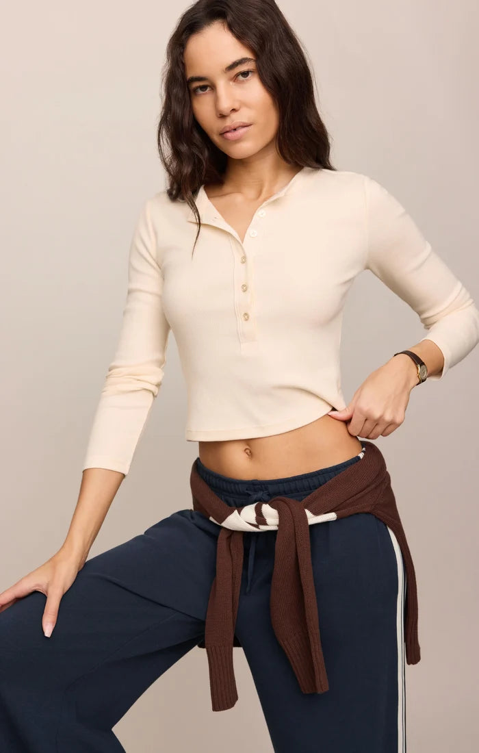 A woman modeling a cream-colored, long-sleeve cropped henley top with a button placket, paired with navy blue pants and a brown tie-waist sweater, against a solid light beige background.
