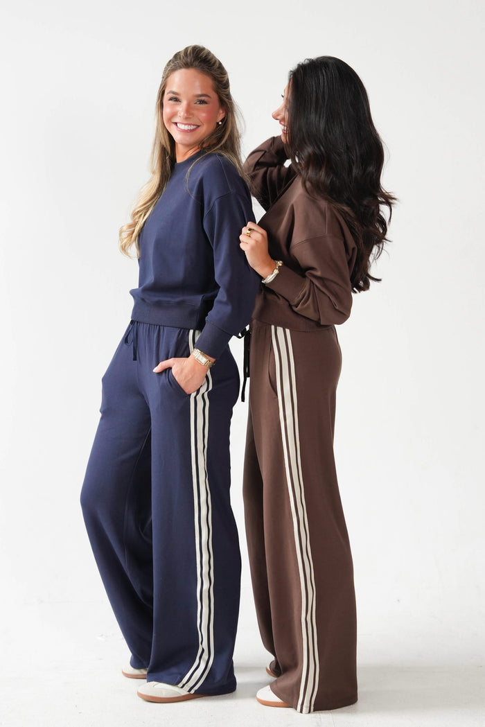 Two women modeling matching lounge sets in a studio setting. The woman on the left wears a navy blue sweatshirt and matching wide-leg pants with white side stripes.