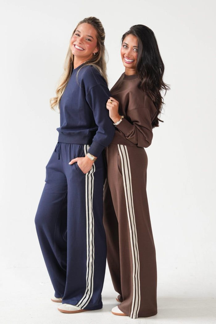 Two women modeling matching lounge sets in navy blue and brown, featuring long-sleeve sweatshirts and wide-leg pants with white side stripes, against a plain white studio background.