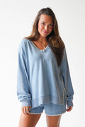 A woman modeling a light blue long-sleeve sweatshirt and matching shorts set with decorative silver trim details, shot against a plain white studio background.