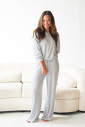 A woman modeling a matching long-sleeve top and wide-leg pants loungewear set with thin gray horizontal stripes on a white background, standing in front of a cream-colored sofa.