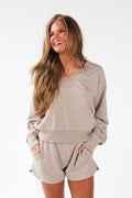 A woman wearing a matching beige lounge set consisting of a long-sleeve V-neck sweatshirt and shorts, smiling against a plain white background.