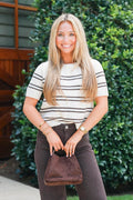 A smiling blonde woman stands outdoors in front of a green hedge, wearing a cream short-sleeve knit top with thin dark horizontal stripes, dark brown pants, and holding a brown suede handbag.