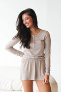 A woman modeling a light taupe ribbed long-sleeve henley romper with a button placket and matching shorts, set against a bright, neutral indoor background.