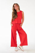 A woman modeling a bright red two-piece lounge set consisting of a sleeveless crewneck top and matching wide-leg pants, paired with white sneakers against a plain white studio background.