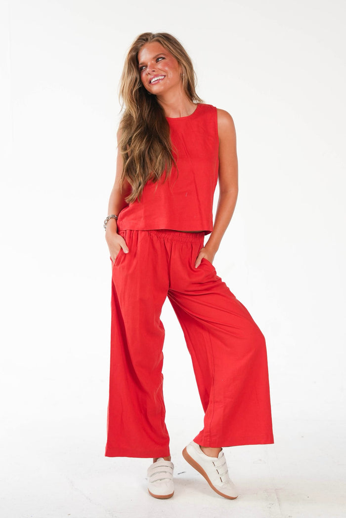 A woman modeling a bright red two-piece lounge set consisting of a sleeveless crewneck top and matching wide-leg pants, paired with white sneakers against a plain white studio background.