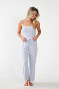 A woman modeling a light blue pajama set consisting of a camisole top with lace trim and matching drawstring pants, standing against a plain white studio background.